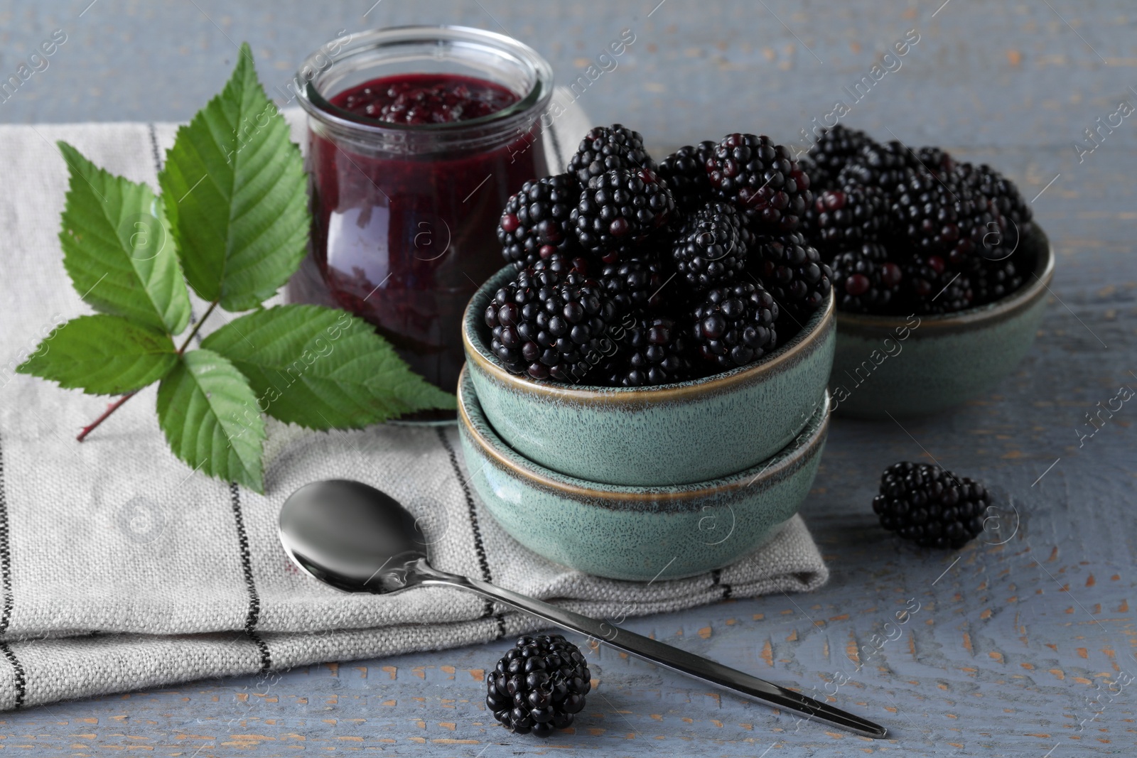 Photo of Fresh ripe blackberries, tasty jam and leaves on light grey wooden table