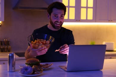 Man eating chips while using laptop in kitchen at night. Bad habit