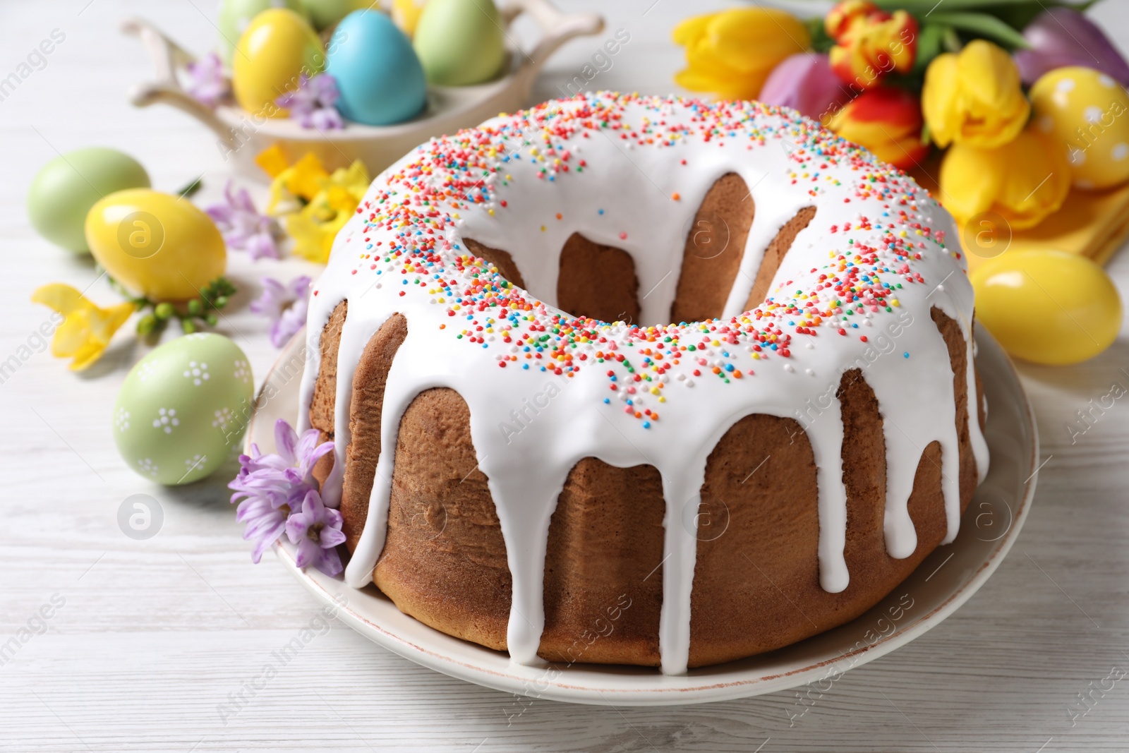 Photo of Glazed Easter cake with sprinkles, painted eggs and flowers on white wooden table, closeup