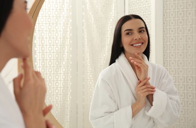 Beautiful young woman in bathrobe near mirror indoors