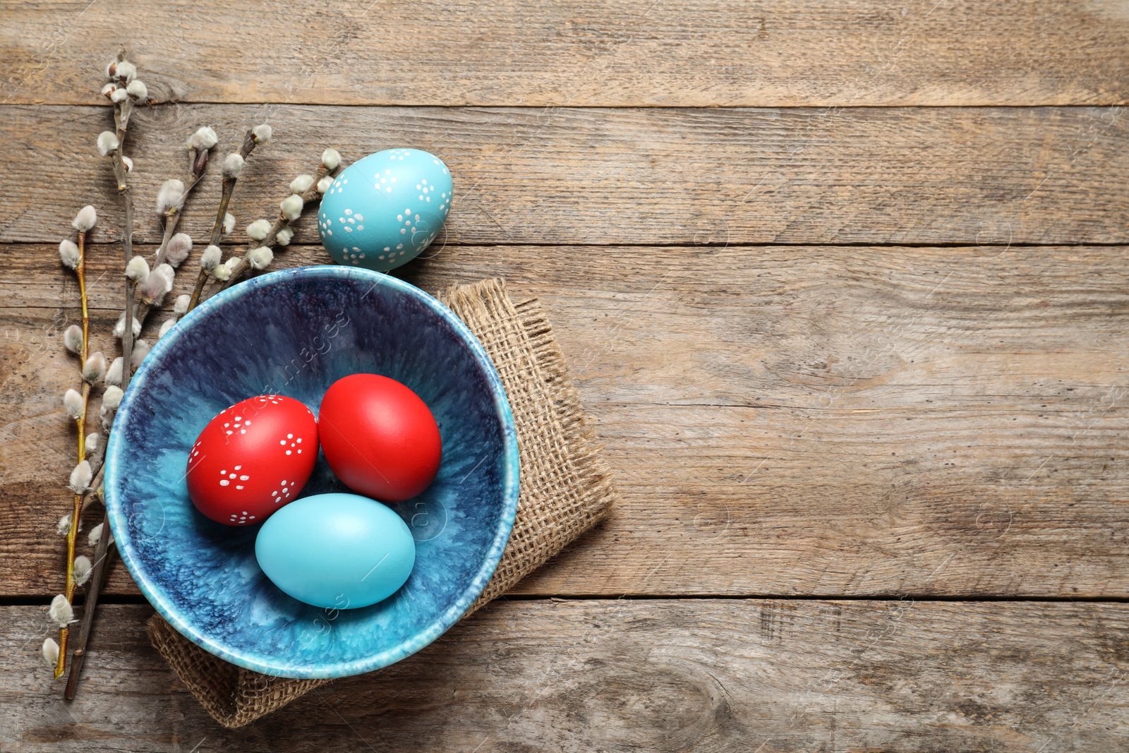 Photo of Flat lay composition of bowl with painted Easter eggs and pussy willow on wooden table, space for text