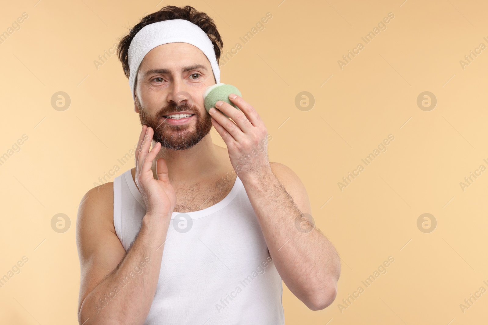 Photo of Man with headband washing his face using sponge on beige background, space for text