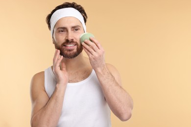 Man with headband washing his face using sponge on beige background, space for text