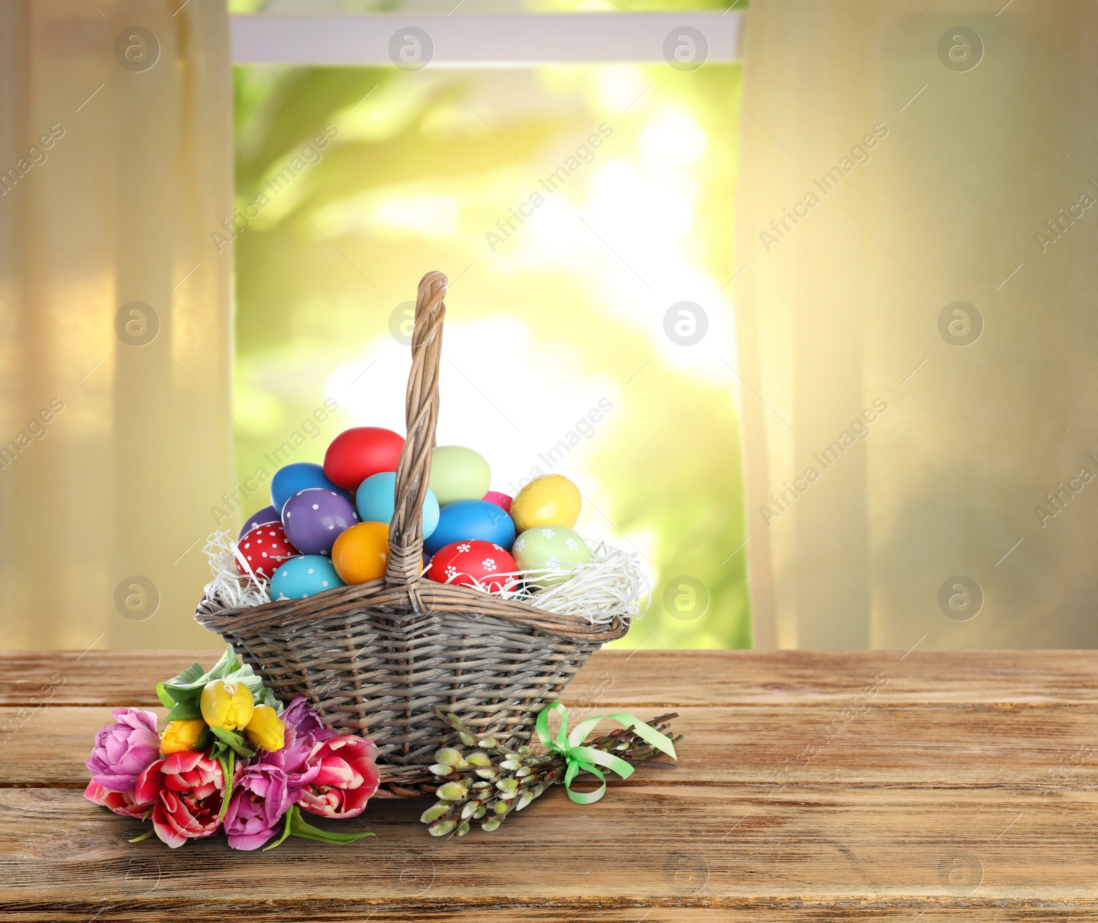 Image of Wicker basket with bright painted Easter eggs and spring flowers on wooden table indoors