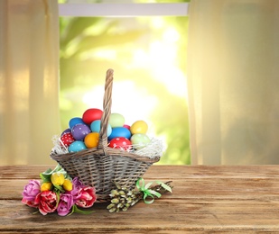 Image of Wicker basket with bright painted Easter eggs and spring flowers on wooden table indoors