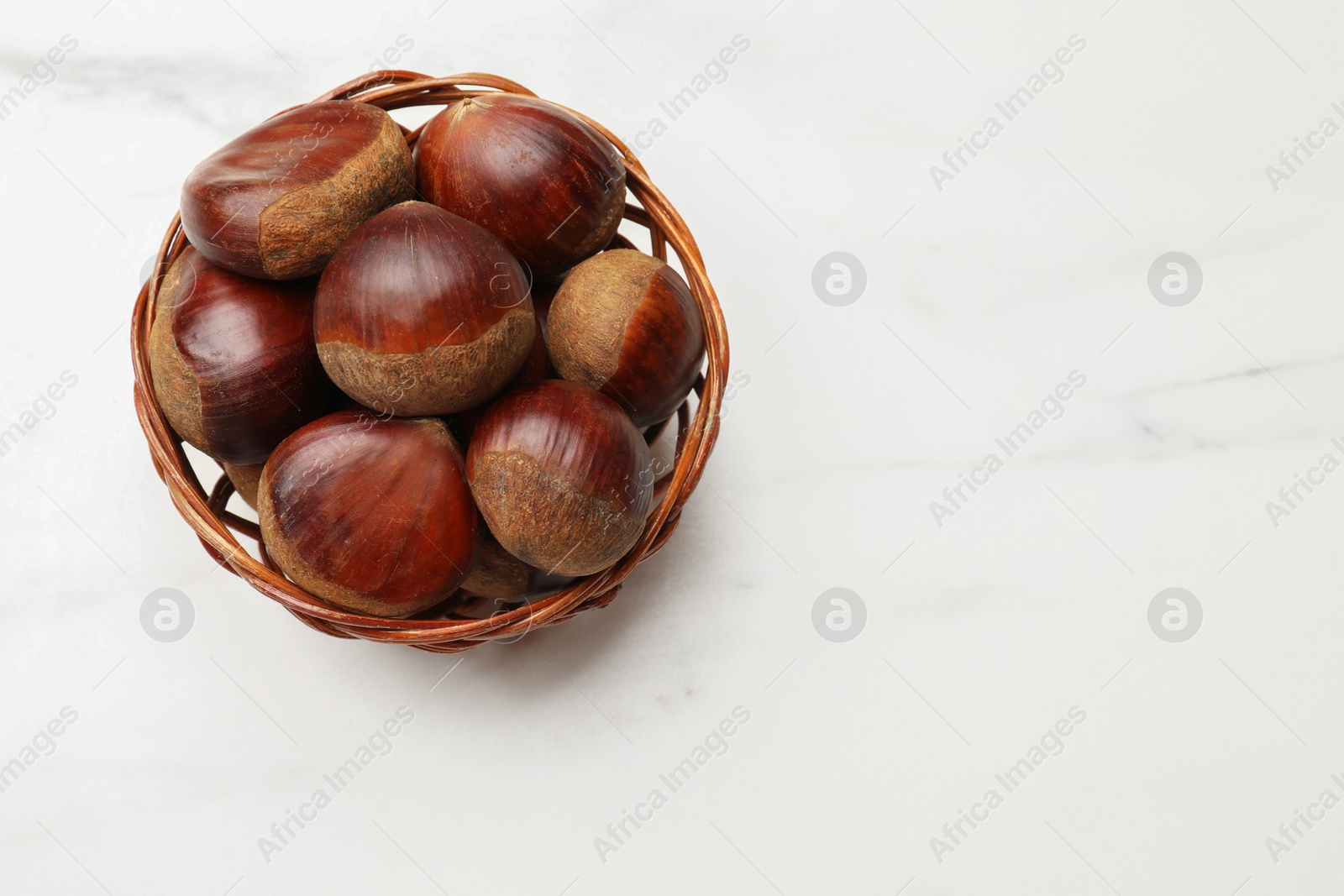 Photo of Sweet fresh edible chestnuts in wicker bowl on white marble table, top view. Space for text