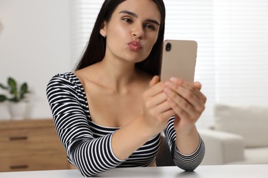 Young woman having video chat via smartphone and sending air kiss at table in room