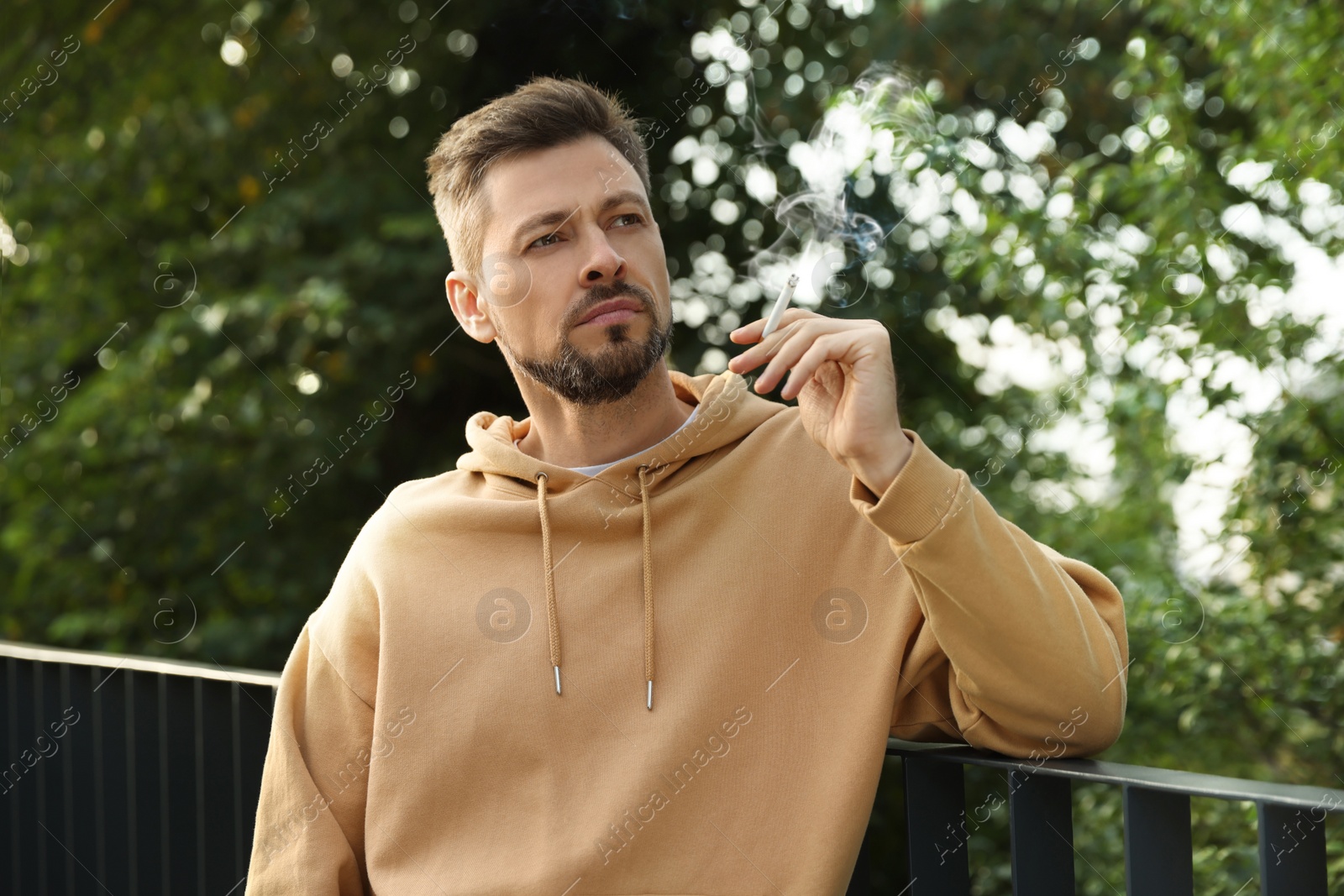 Photo of Man smoking cigarette near railing outdoors. Bad habit