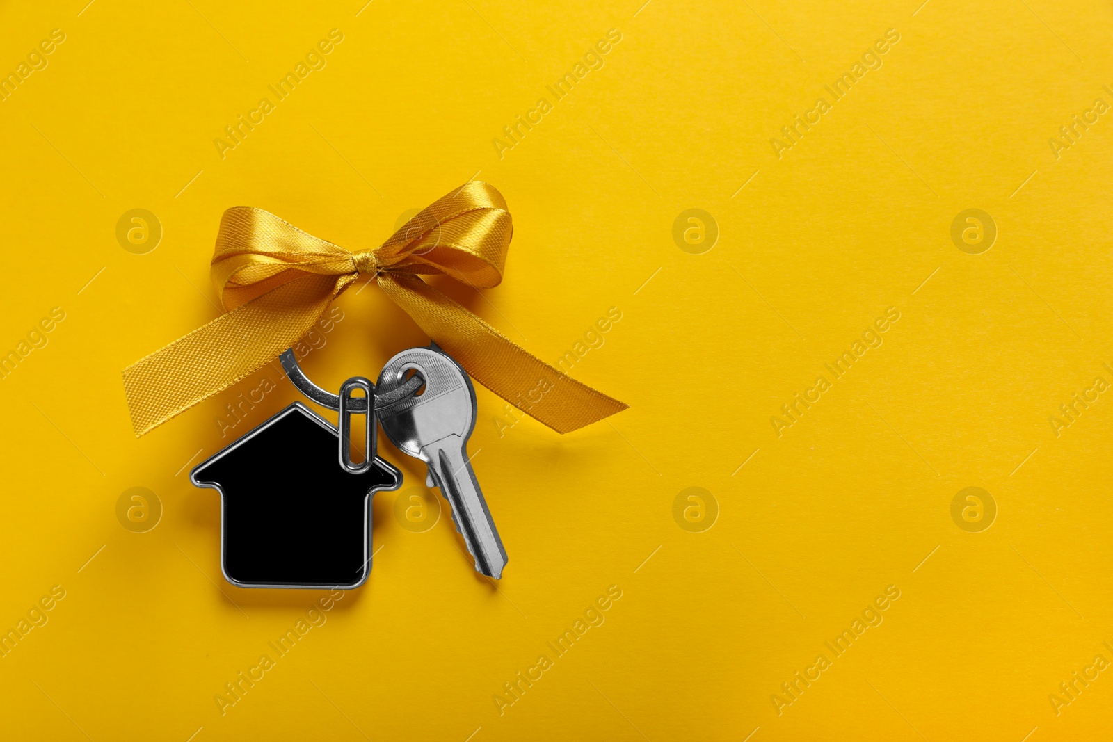 Photo of Key with trinket in shape of house and bow on yellow background, top view. Space for text. Housewarming party
