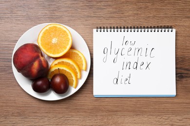 Notebook with words Low Glycemic Index Diet and plate of fresh fruits on wooden table, flat lay