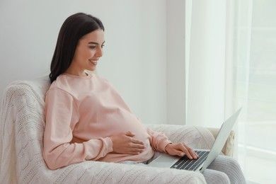 Pregnant young woman with laptop at home, space for text