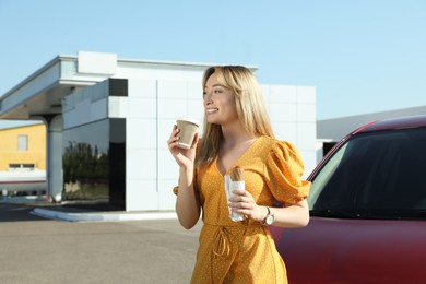 Photo of Beautiful young woman with hot dog drinking coffee near car at gas station