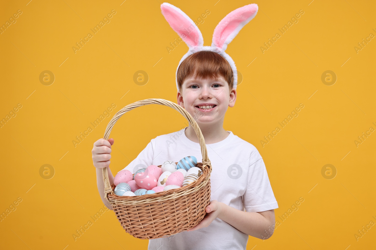 Photo of Easter celebration. Cute little boy with bunny ears and wicker basket full of painted eggs on orange background