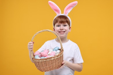 Photo of Easter celebration. Cute little boy with bunny ears and wicker basket full of painted eggs on orange background