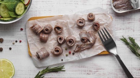 Photo of Canned anchovy fillets, rosemary and lime on white wooden table, flat lay