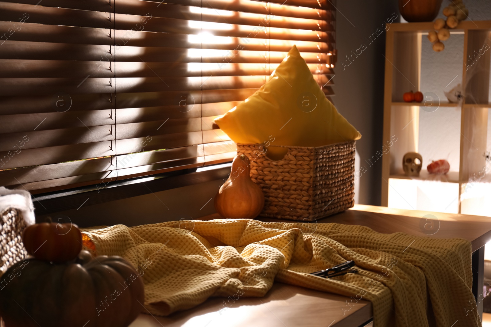 Photo of Yellow blanket, wicker basket and pumpkin on wooden table near window indoors. Halloween celebration