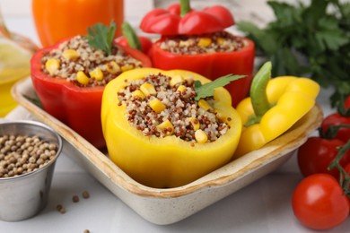 Photo of Quinoa stuffed bell peppers in baking dish, parsley, tomatoes and peppercorns on white tiled table, closeup