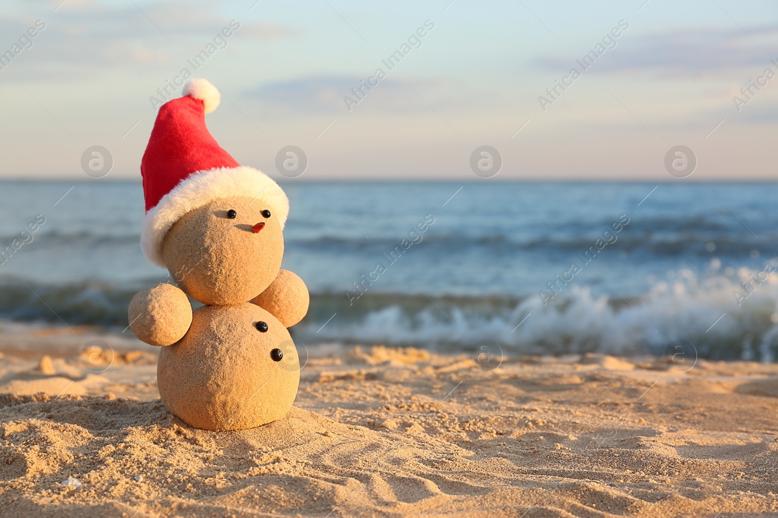 Photo of Snowman made of sand with Santa hat on beach near sea, space for text. Christmas vacation