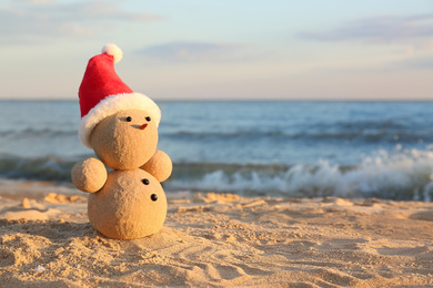 Photo of Snowman made of sand with Santa hat on beach near sea, space for text. Christmas vacation