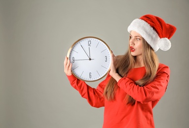 Photo of Young beautiful woman in Santa hat holding big clock on grey background. Christmas celebration