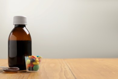 Bottle of syrup, dosing spoon and measuring cup with pills on wooden table against white background, space for text. Cold medicine