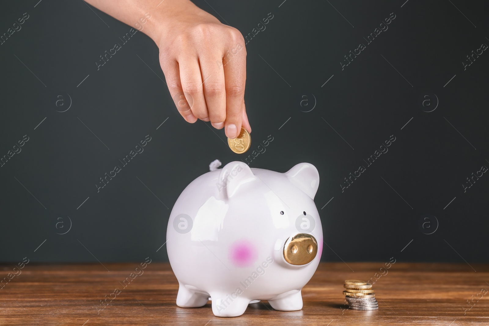 Photo of Woman putting coin into piggy bank on wooden table against dark background. Pension planning