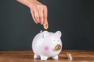 Photo of Woman putting coin into piggy bank on wooden table against dark background. Pension planning