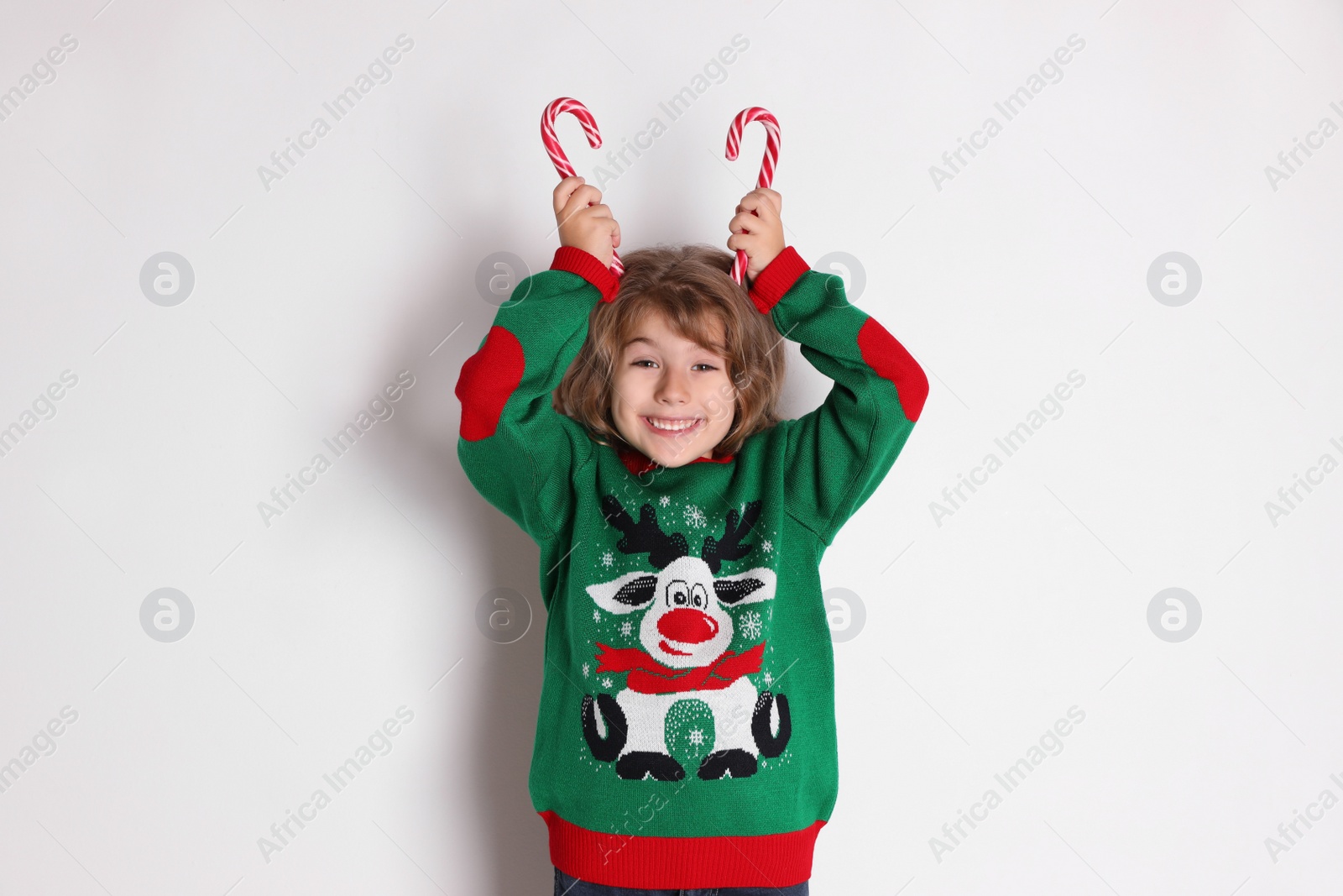 Photo of Cute little girl in Christmas sweater holding sweet candy canes near head against white background