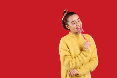 Photo of Young woman in yellow sweater and festive headband eating candy cane on red background, space for text