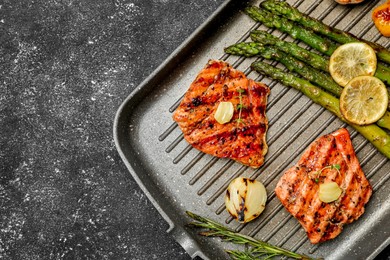 Photo of Frying pan with tasty grilled salmon, lemon and asparagus on black table, top view. Space for text