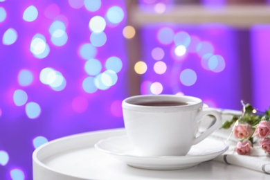 Photo of Cup of hot drink and flowers on table against blurred lights, space for text