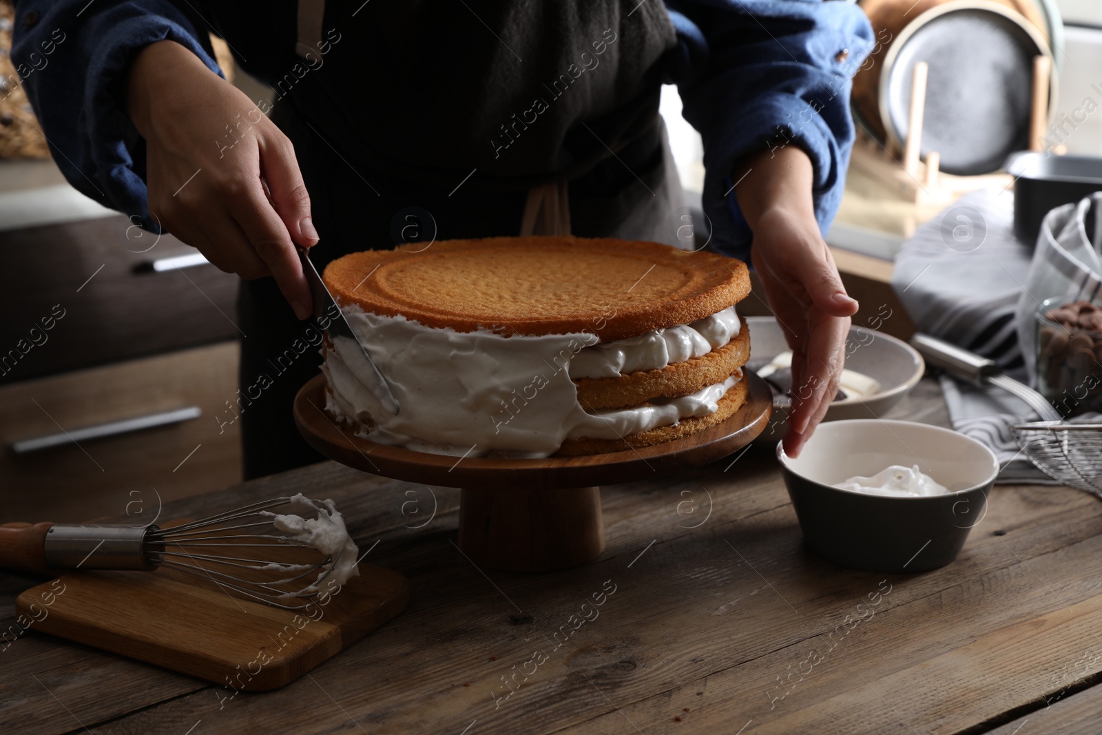 Photo of Woman smearing sides of sponge cake with cream at wooden table, closeup