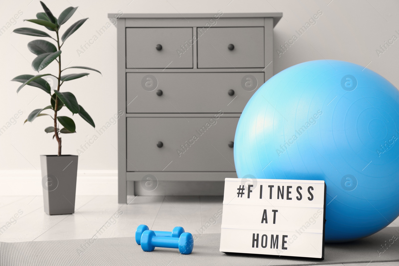 Photo of Sport equipment and lightbox with hashtag FITNESS AT HOME on floor indoors. Message to promote self-isolation during COVID‑19 pandemic