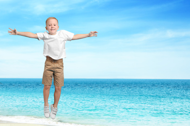 Image of Cute school boy jumping on beach near sea, space for text. Summer holidays