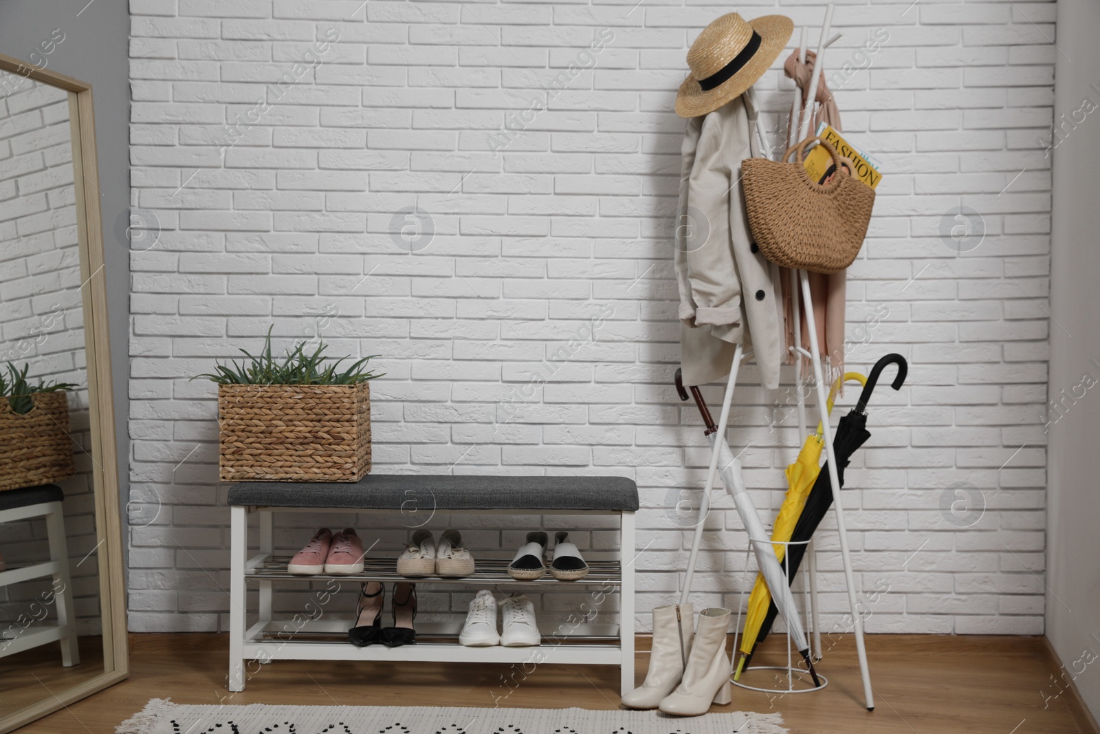 Photo of Stylish hallway interior with coat rack, shoe storage bench and mirror near white brick wall