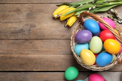 Colorful Easter eggs in wicker basket and tulips on wooden table, flat lay. Space for text