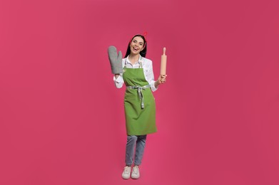 Young housewife in oven glove holding rolling pin on pink background