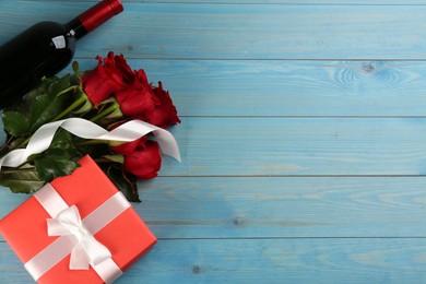Beautiful red roses, gift box and bottle of wine on light blue wooden background, flat lay with space for text. Valentine's Day celebration