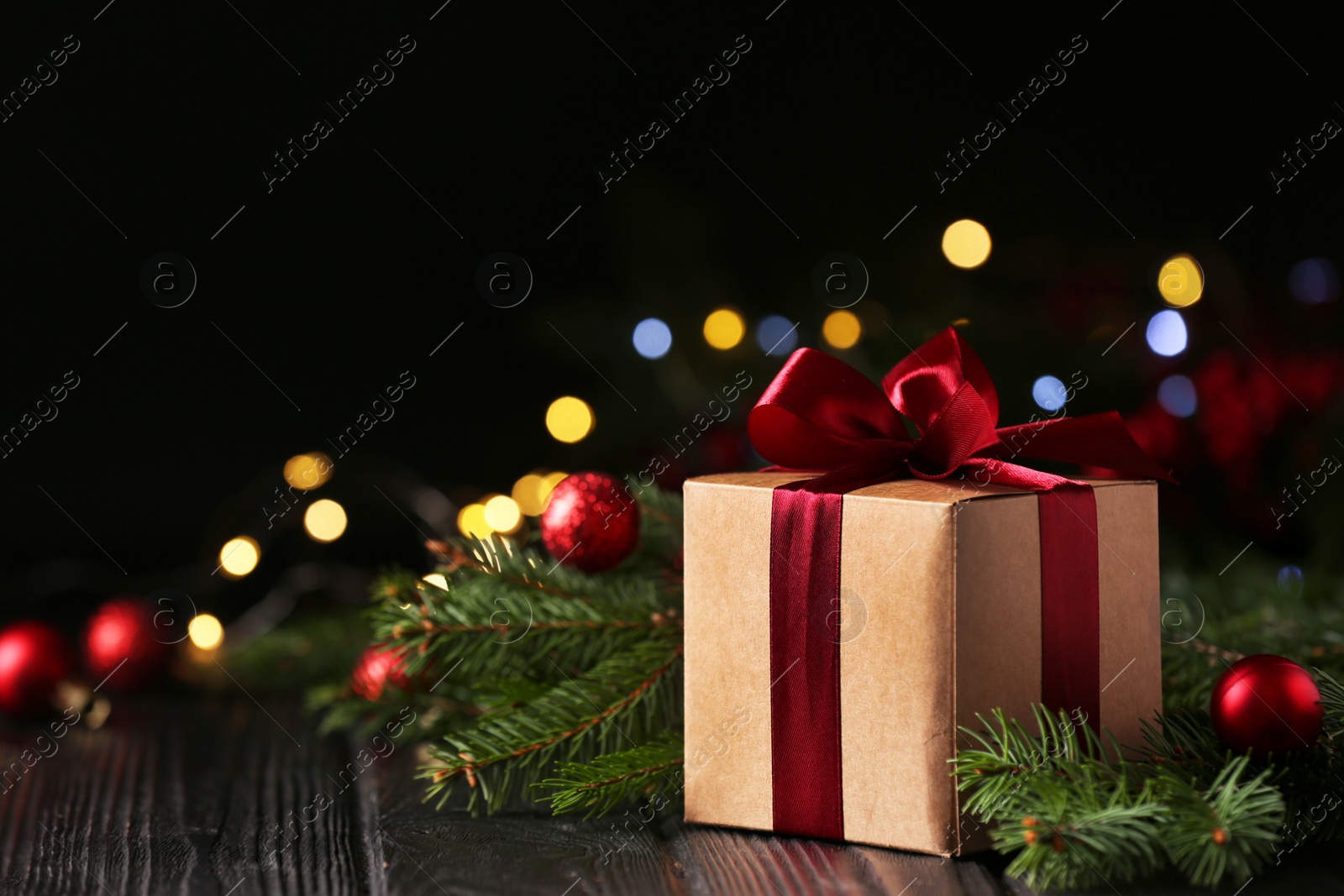 Photo of Beautiful gift box, fir tree branches and Christmas decor on wooden table against blurred festive lights, space for text