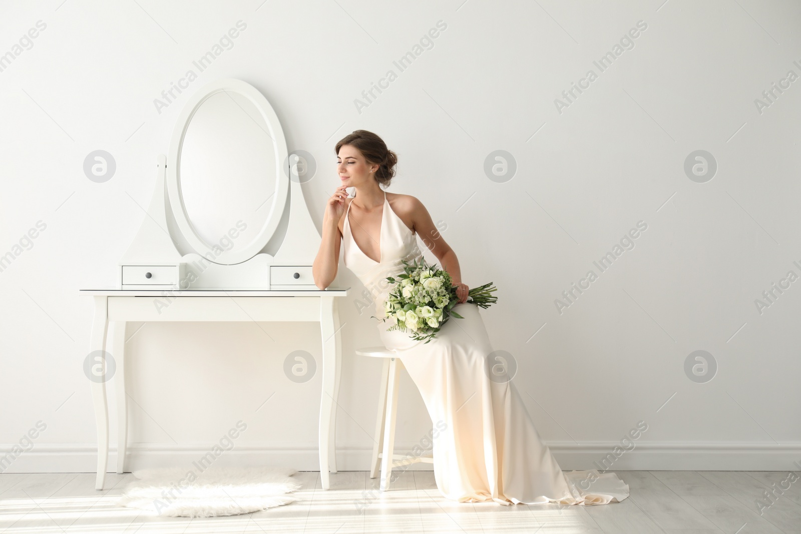 Photo of Young bride in wedding dress with beautiful bouquet near mirror indoors