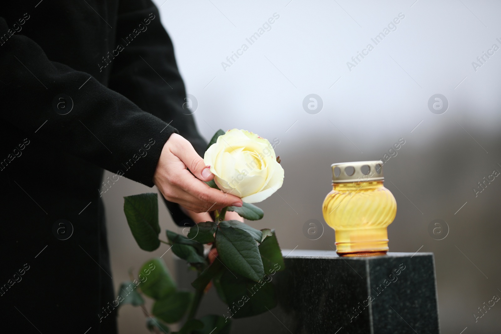 Photo of Woman holding white rose near black granite tombstone with candle outdoors, closeup. Funeral ceremony