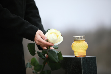 Photo of Woman holding white rose near black granite tombstone with candle outdoors, closeup. Funeral ceremony