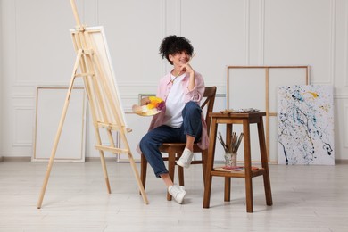 Photo of Young woman holding brush and artist`s palette near easel with canvas