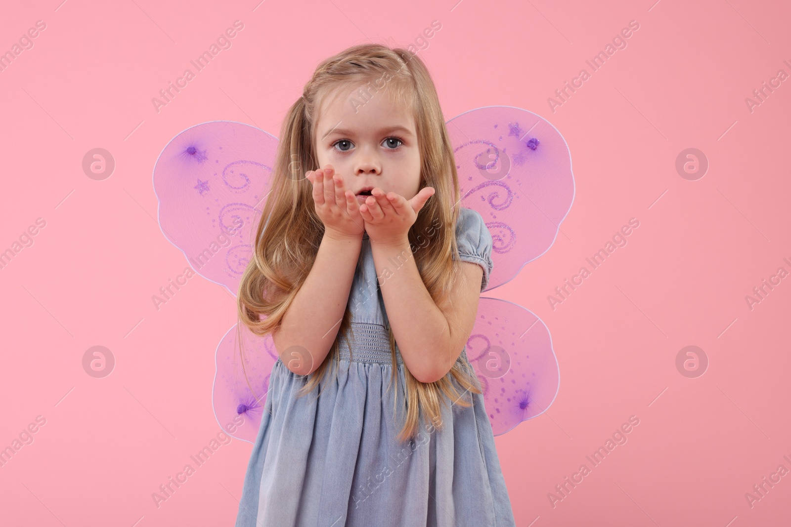 Photo of Cute little girl in fairy costume with violet wings blowing kiss on pink background