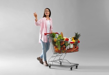 Young woman with shopping cart full of groceries on grey background