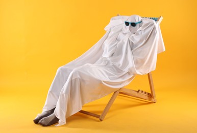 Photo of Person in ghost costume and sunglasses relaxing on deckchair against yellow background