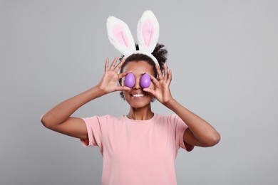 Photo of Happy African American woman in bunny ears headband covering eyes with Easter eggs on gray background