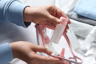 Photo of Cosmetic travel kit. Woman putting small bottle with personal care product into plastic bag on bed, closeup