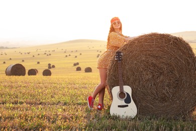 Photo of Happy hippie woman with guitar near hay bale in field, space for text