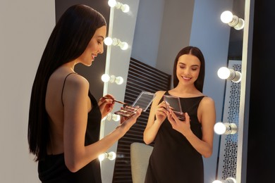 Photo of Beautiful young woman in elegant dress with eyeshadow palette and brush near mirror indoors